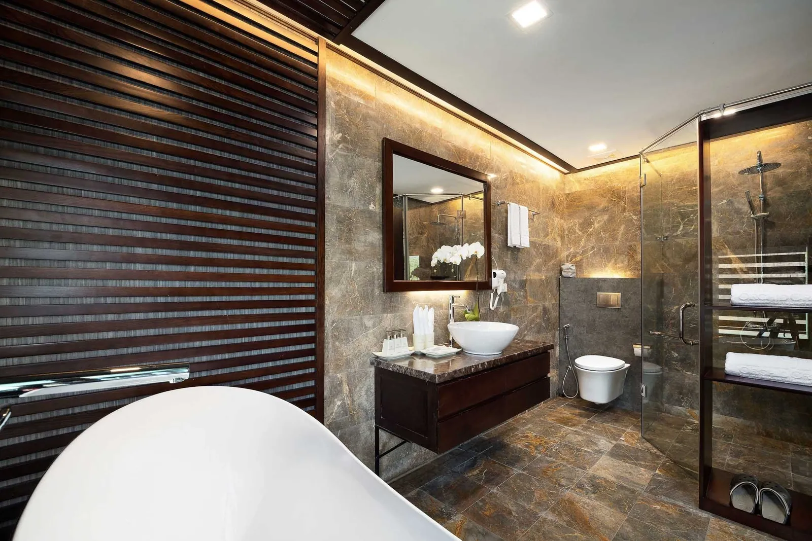 A marble-tiled ensuite bathroom with a white freestanding bathtub, a vessel sink on a dark wood vanity, a framed mirror, a wall-hung toilet, and a glass shower enclosure with towels and slippers.