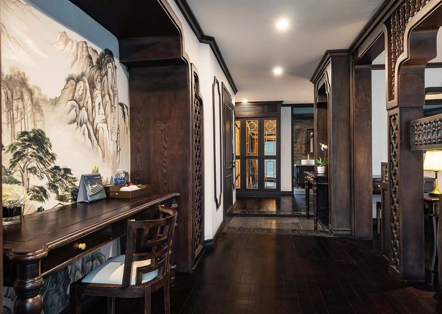 Interior of a dark-wood suite with a wooden writing desk and chair, an ink-style mountain landscape mural, carved lattice wood partitions, and a hallway leading to a glass-paneled wardrobe and bathroom.