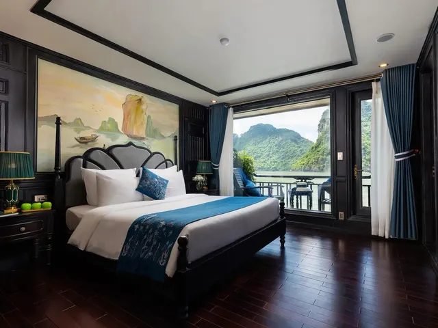 A cruise boat cabin with a bed, dark wood paneling, and a balcony door opening to a view of karst limestone mountains and calm water.
