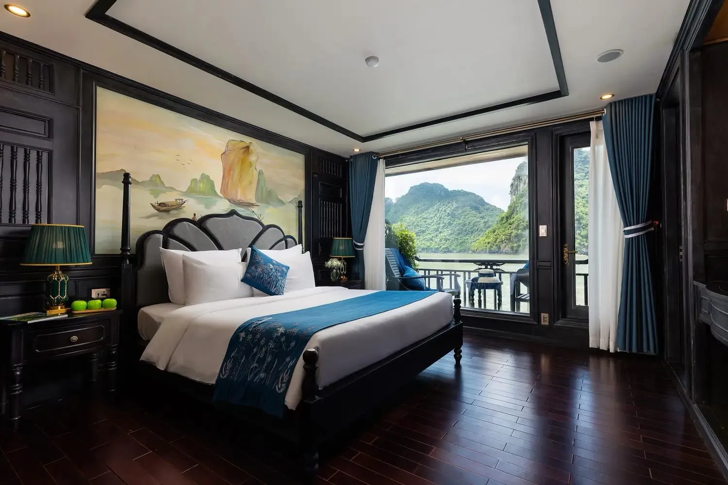 A cruise boat cabin with a bed, dark wood paneling, and a balcony door opening to a view of karst limestone mountains and calm water.