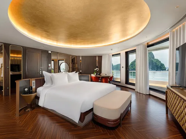 A luxury cruise cabin with a king bed, gold coffered ceiling, herringbone wood floor, and large windows revealing karst limestone islands on the water outside.