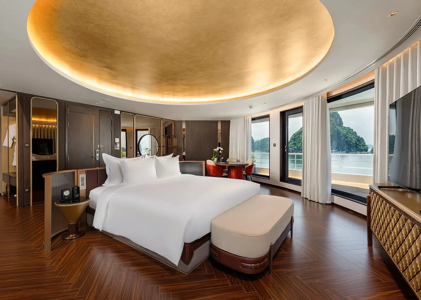 A luxury cruise cabin with a king bed, gold coffered ceiling, herringbone wood floor, and large windows revealing karst limestone islands on the water outside.