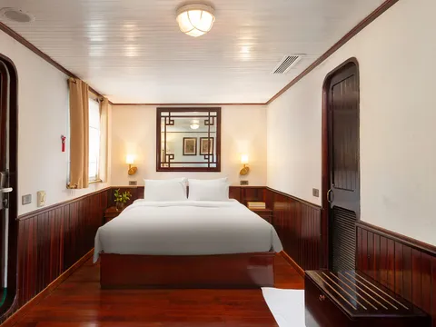 A cabin with a double bed in white linens, dark red-brown wood paneling and floor, a latticed mirror, wall lamps, a window with tan curtains, and louvered wooden doors on both sides.