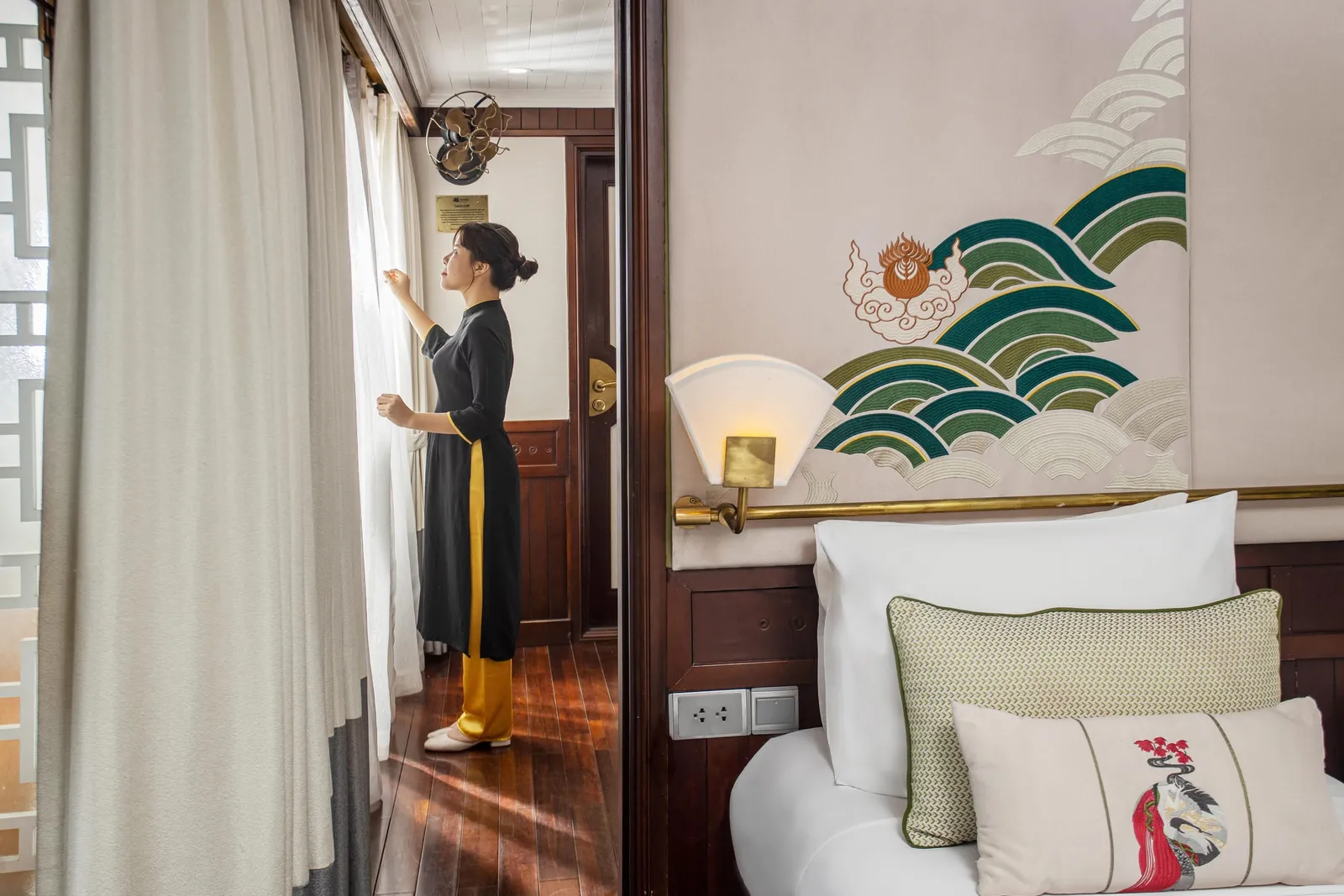 A staff member in traditional dress opens curtains in a cabin with a bed, decorative wave mural, and wooden furnishings.