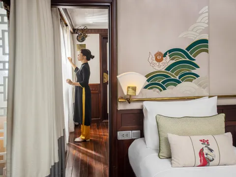 A staff member in traditional dress opens curtains in a cabin with a bed, decorative wave mural, and wooden furnishings.