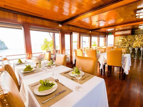 A formally set dining room inside a cruise boat featuring warm wood panelling, white-clothed tables with wine glasses and folded green napkins, leather chairs, and limestone karst formations visible through large windows overlooking the water.