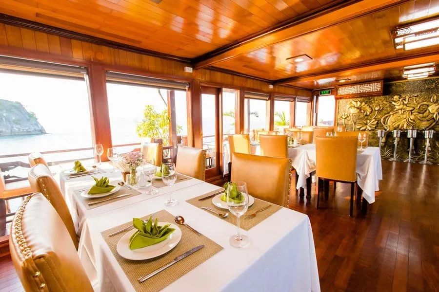 A formally set dining room inside a cruise boat featuring warm wood panelling, white-clothed tables with wine glasses and folded green napkins, leather chairs, and limestone karst formations visible through large windows overlooking the water.