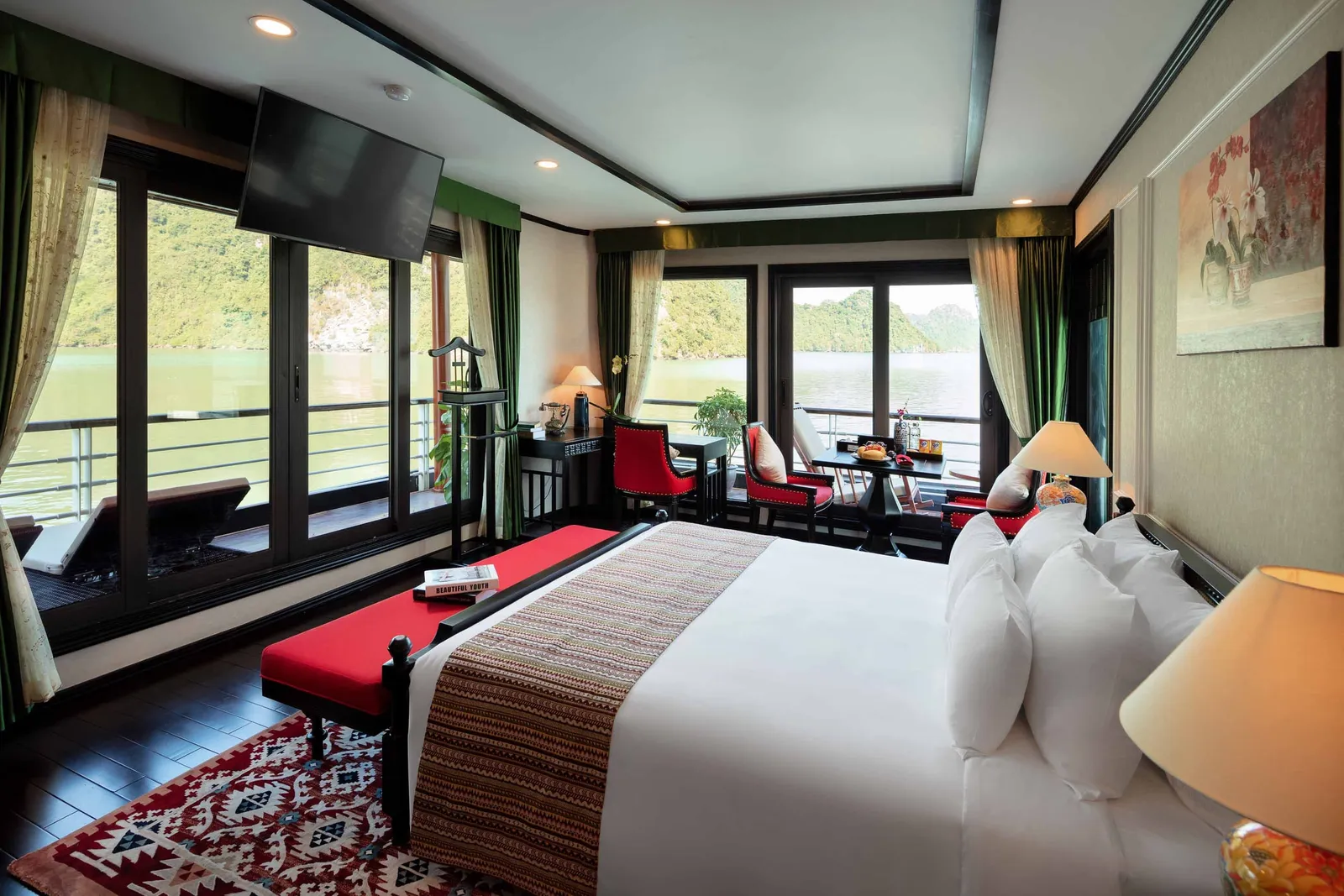 A cruise cabin interior with a bed, seating area, and large windows overlooking calm green water with limestone karst formations in the distance.