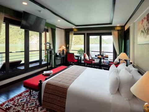 A cruise cabin interior with a bed, seating area, and large windows overlooking calm green water with limestone karst formations in the distance.