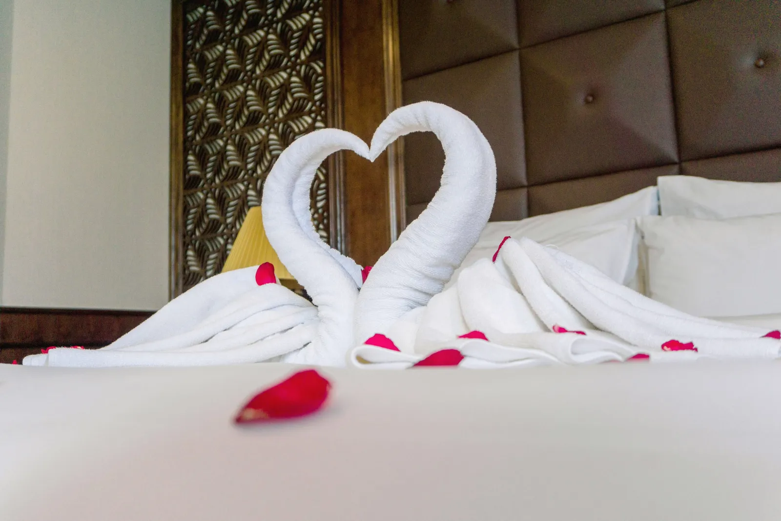 Towels folded into two swans meeting over a heart of rose petals on a white bed, quilted headboard behind