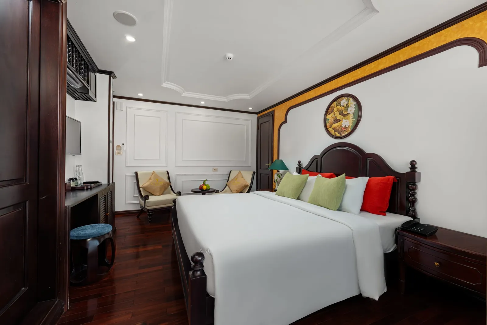 An interior cabin with a bed, dark wood furniture, white walls with yellow trim, and two chairs near a window.