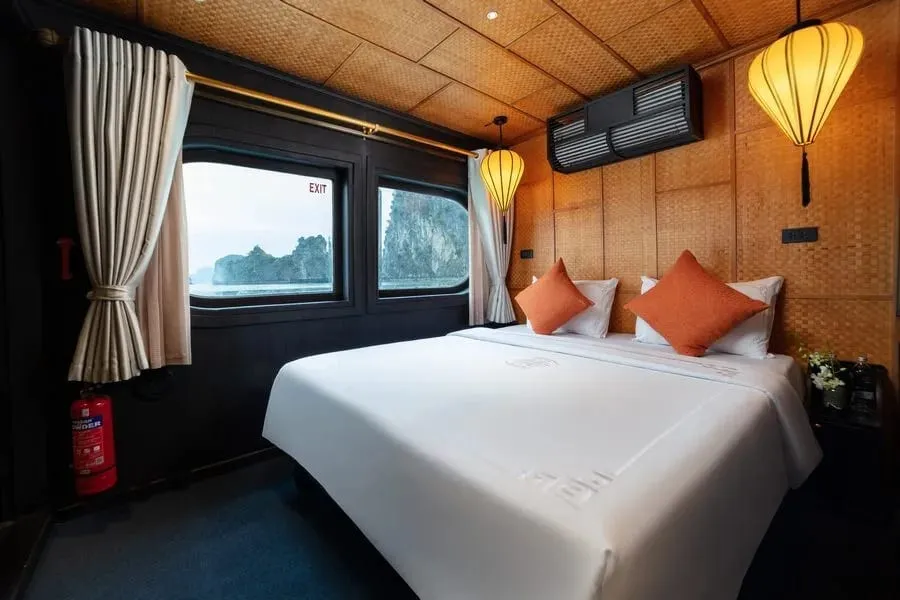 A cabin with a white double bed, orange cushions, woven bamboo walls and silk lanterns, with porthole windows showing limestone karsts over misty water, a fire extinguisher and an EXIT sign, and a subtle embroidered pattern on the bed linen.