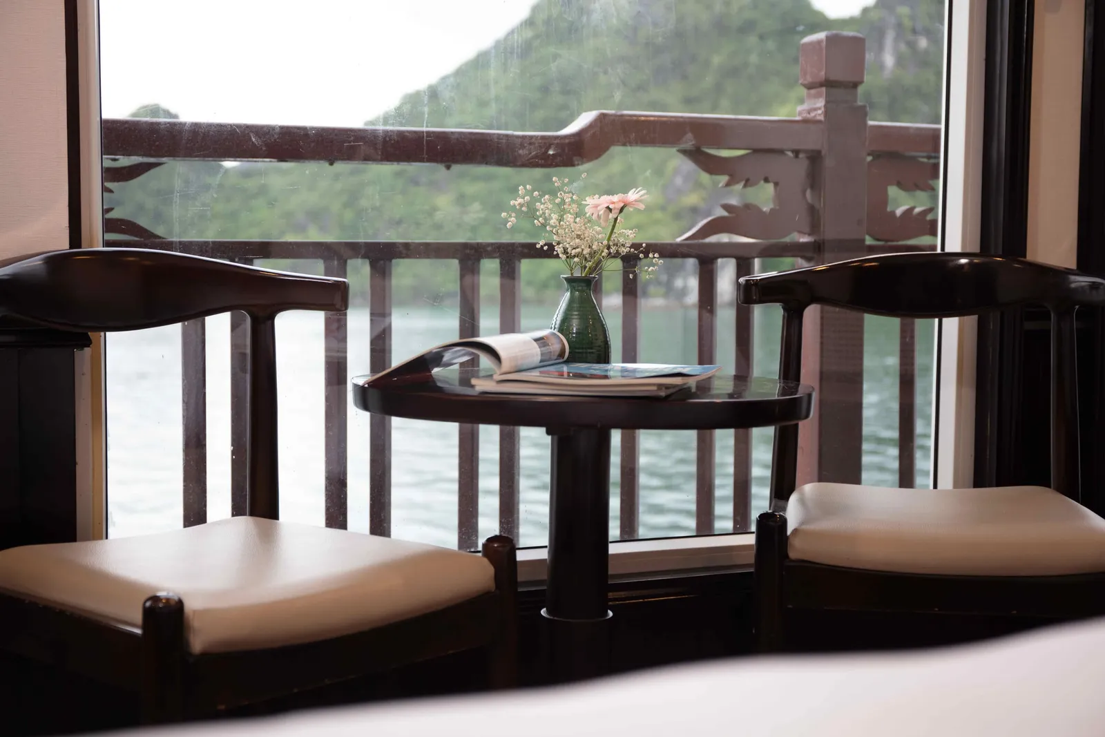 Two dark wooden chairs with cream cushions flank a small round table holding an open magazine and a green vase of flowers, in front of a window overlooking a wooden balcony railing, calm water and a green karst hill.