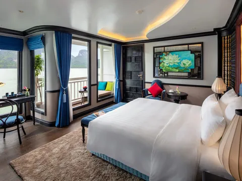 A cabin with a large white bed, blue curtains, a desk and a private balcony, with floor-to-ceiling windows looking onto calm water and limestone karst islands.