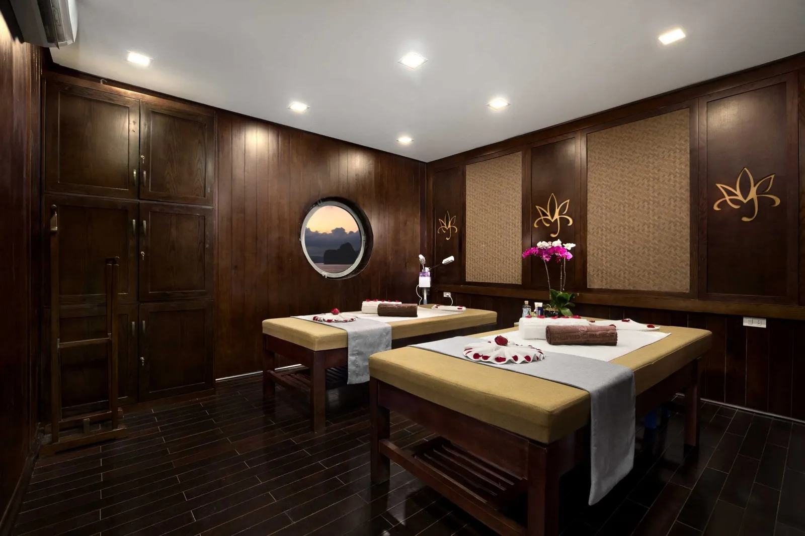 A spa or massage room interior with two massage tables, dark wood paneling, a circular porthole window showing sky, and lotus decorative motifs on the walls.