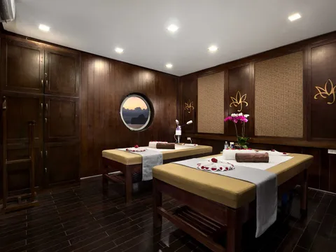 A spa or massage room interior with two massage tables, dark wood paneling, a circular porthole window showing sky, and lotus decorative motifs on the walls.