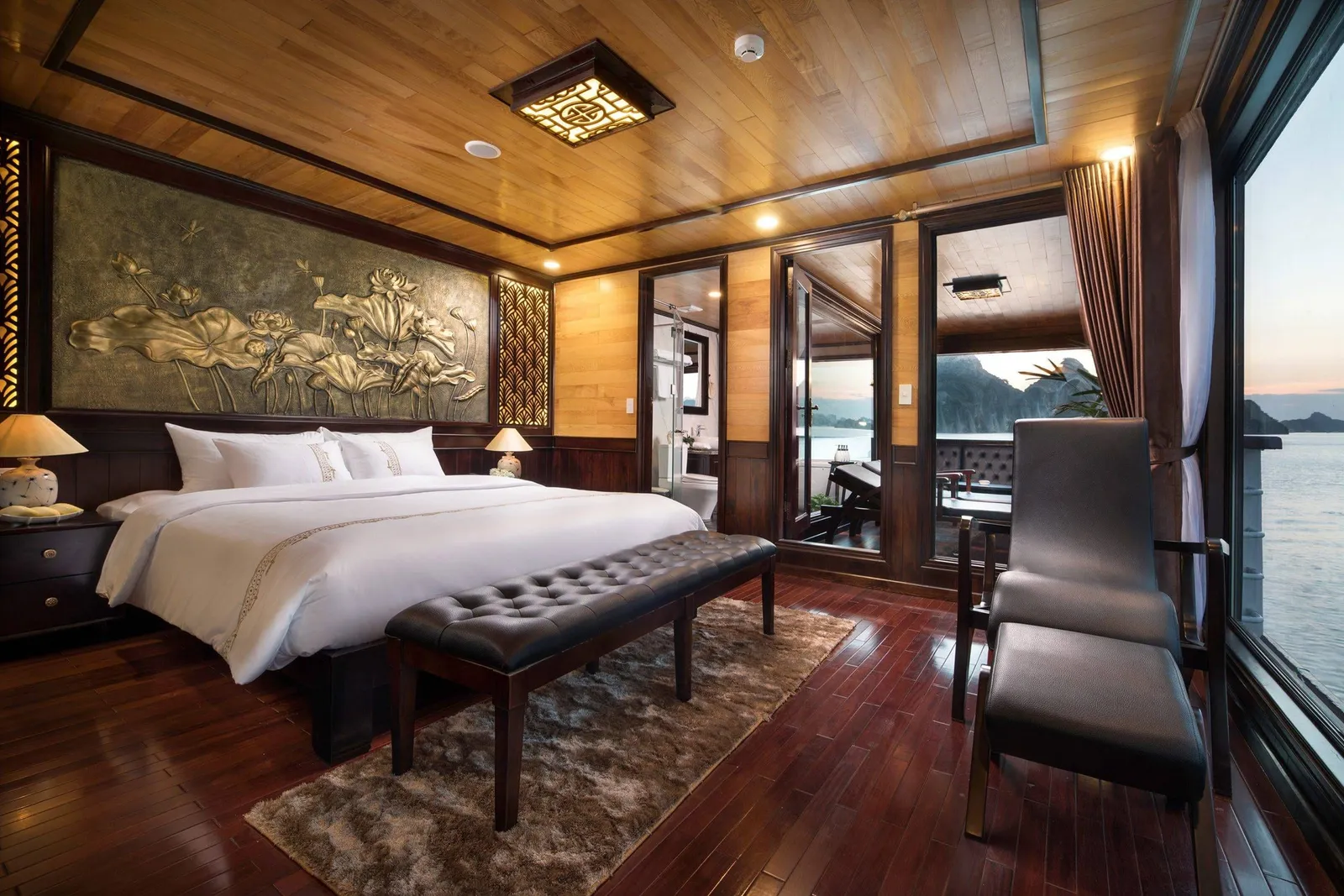 A wood-paneled cruise cabin with a white-bedded king bed, leather bench, lotus relief panel, armchair and ottoman, with a private balcony and a window overlooking limestone karst islands and calm water at dusk.