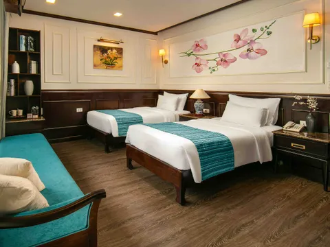 A cruise ship cabin with two twin beds, dark wood furniture, white paneled walls with orchid artwork, teal accents, and warm lighting.