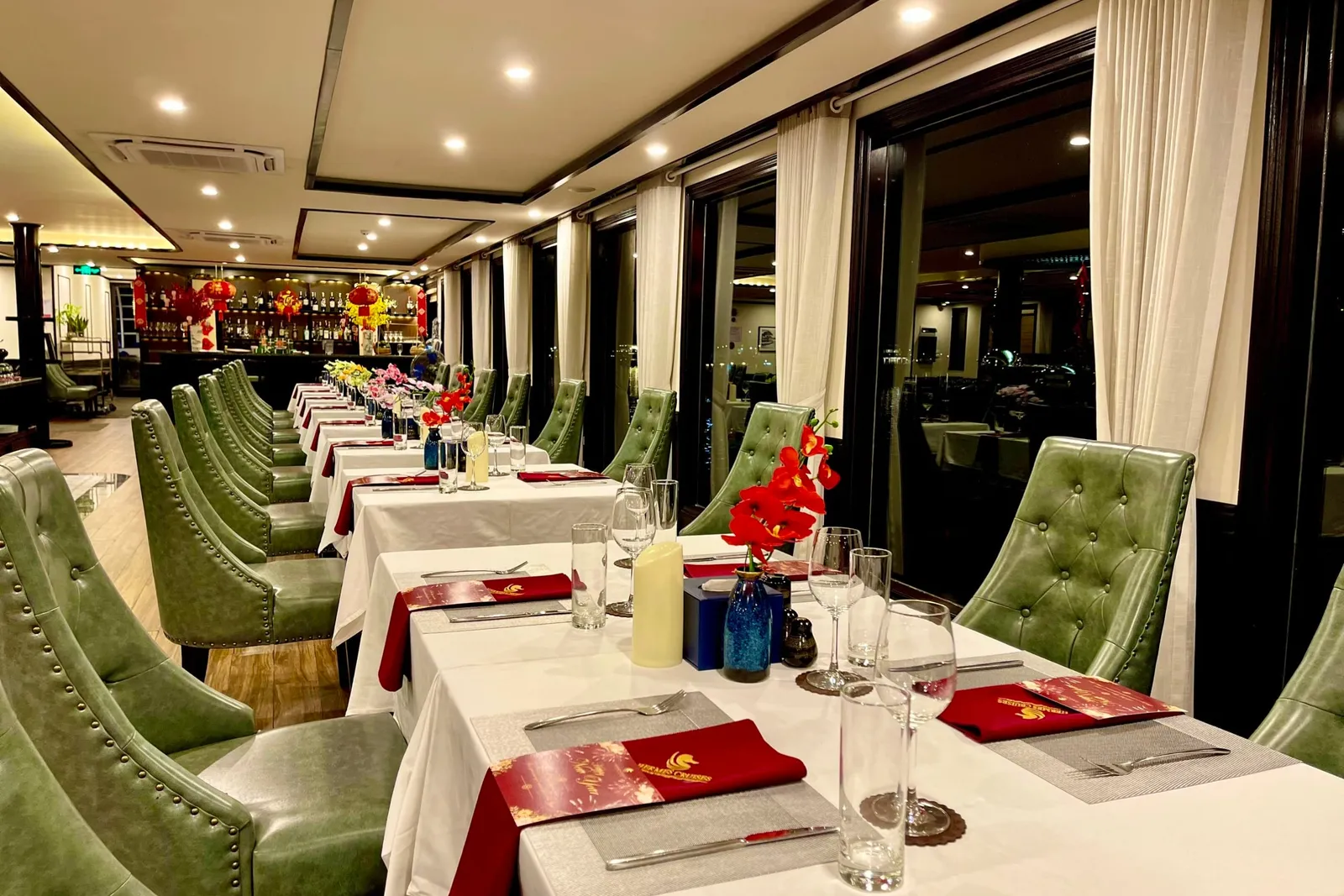 An elegant dining room with green tufted chairs, white-clothed tables set with red menus and napkins, large windows along one side, and red lantern decorations visible in the bar area.