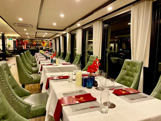 An elegant dining room with green tufted chairs, white-clothed tables set with red menus and napkins, large windows along one side, and red lantern decorations visible in the bar area.