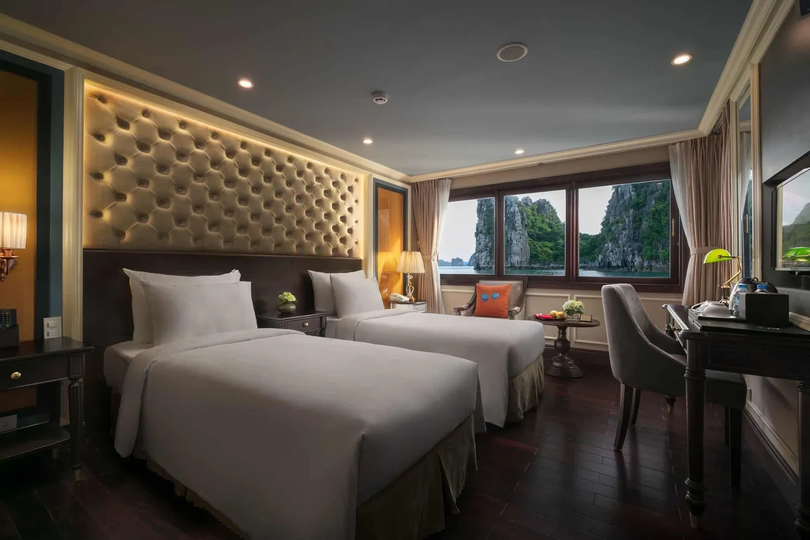 A cruise cabin with two twin beds, a tufted lit headboard, a desk and chair, and windows showing green limestone karsts rising from the water.