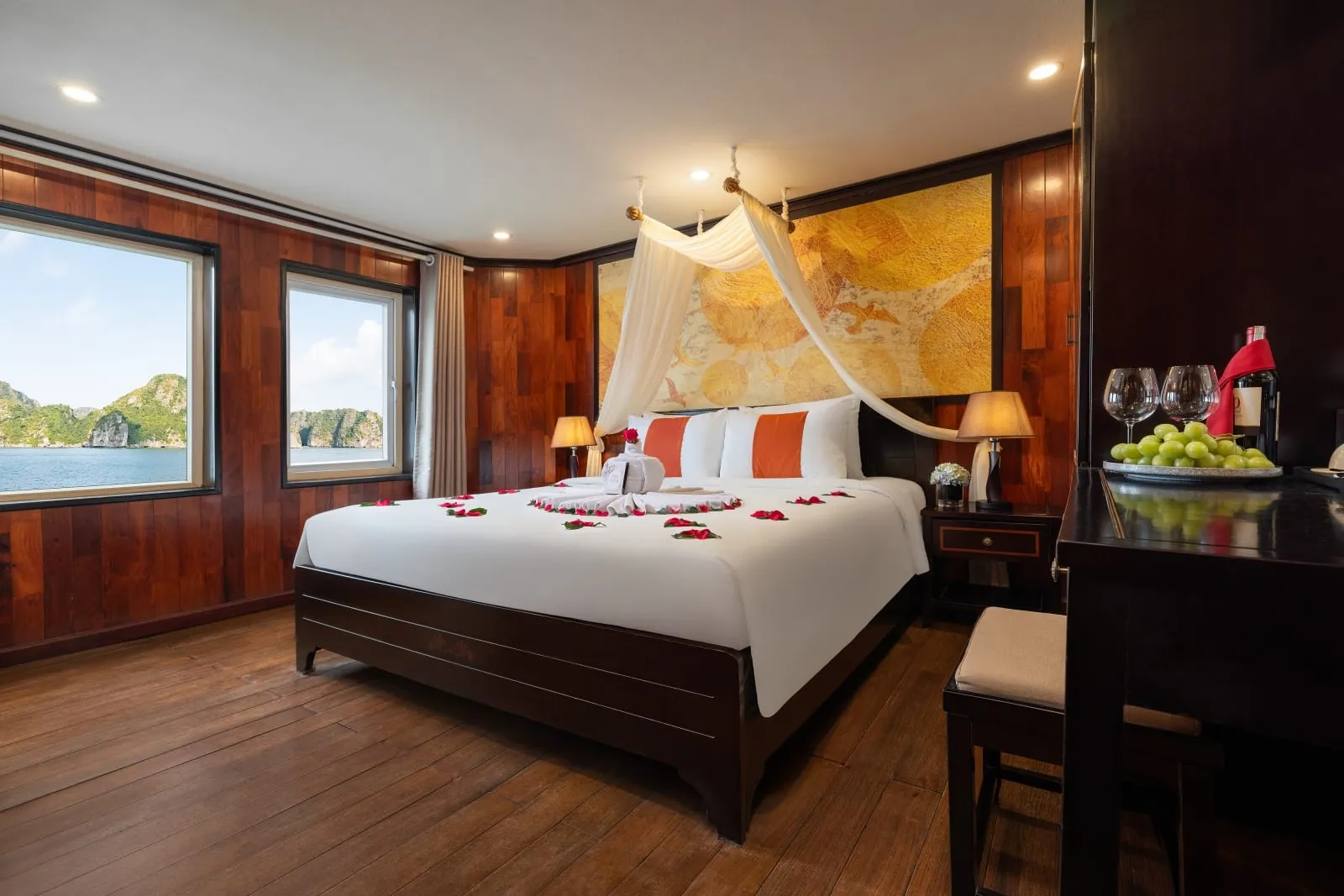 A wooden-paneled cabin with a bed decorated with rose petals, mosquito netting, and windows showing karst limestone formations on water.