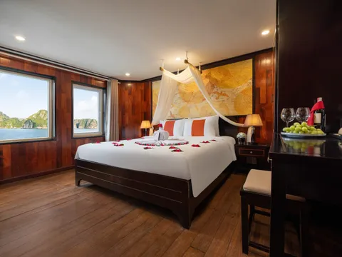 A wooden-paneled cabin with a bed decorated with rose petals, mosquito netting, and windows showing karst limestone formations on water.