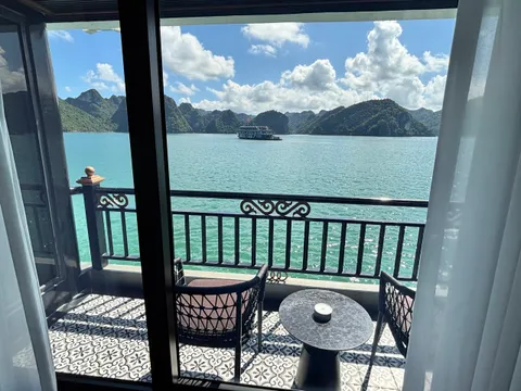 View through open sliding doors from a cabin onto a private balcony with two chairs, a small round table and patterned tile floor, overlooking turquoise water, limestone karst islands and another cruise boat under a partly cloudy sky.