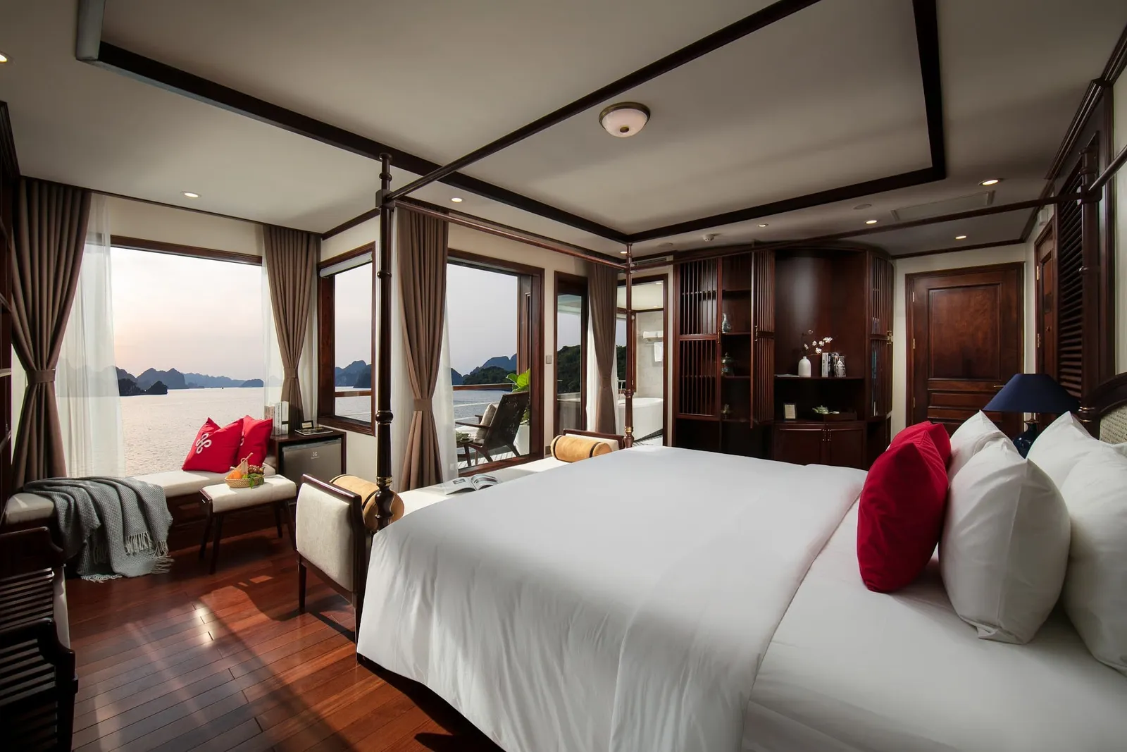 A luxury cabin interior with a white bed, dark wood furnishings, and large windows showing limestone karst islands on calm water.