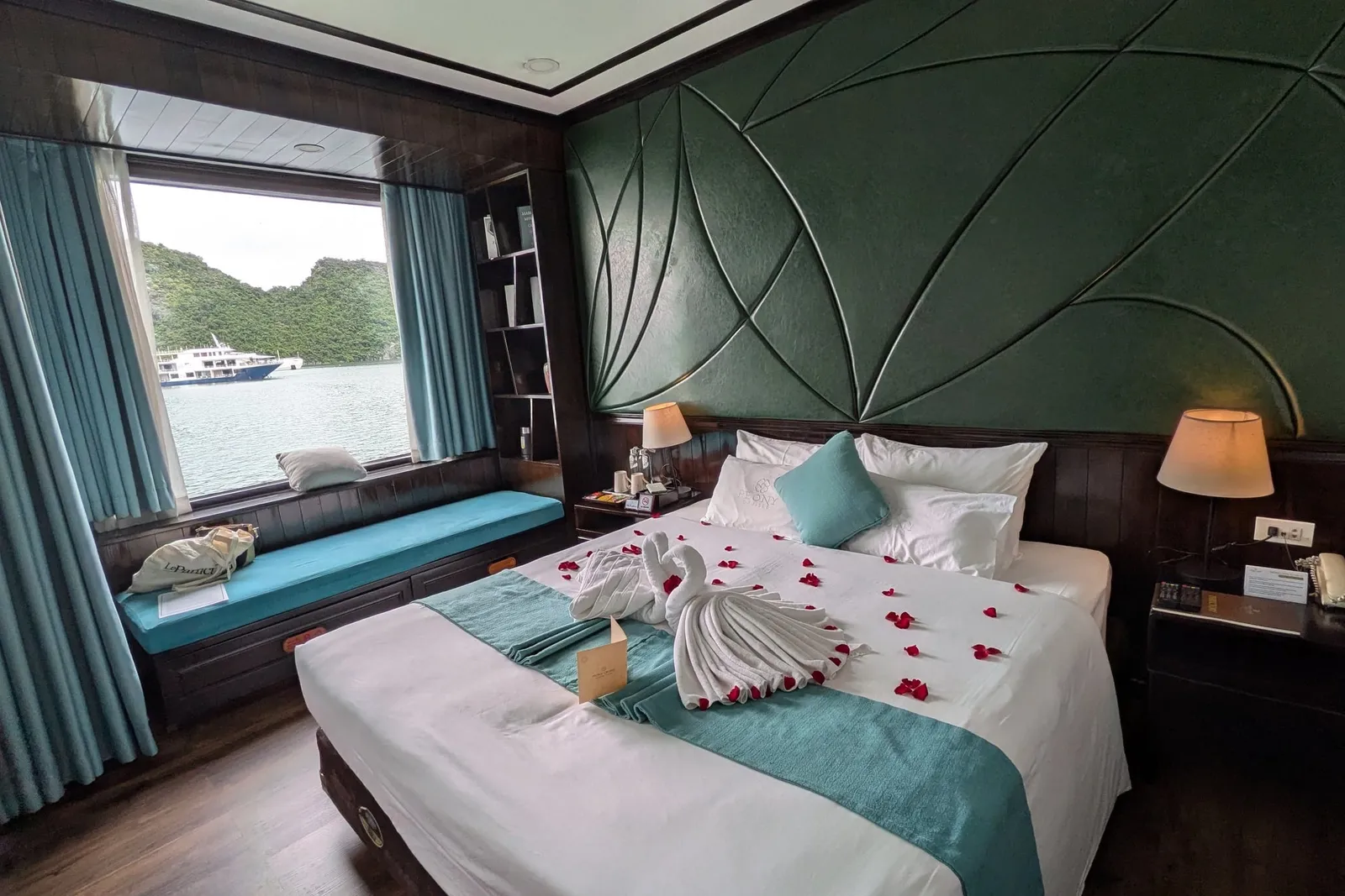 Cabin with a quilted green headboard, towels folded into a swan on the bed and a padded window seat looking onto the bay