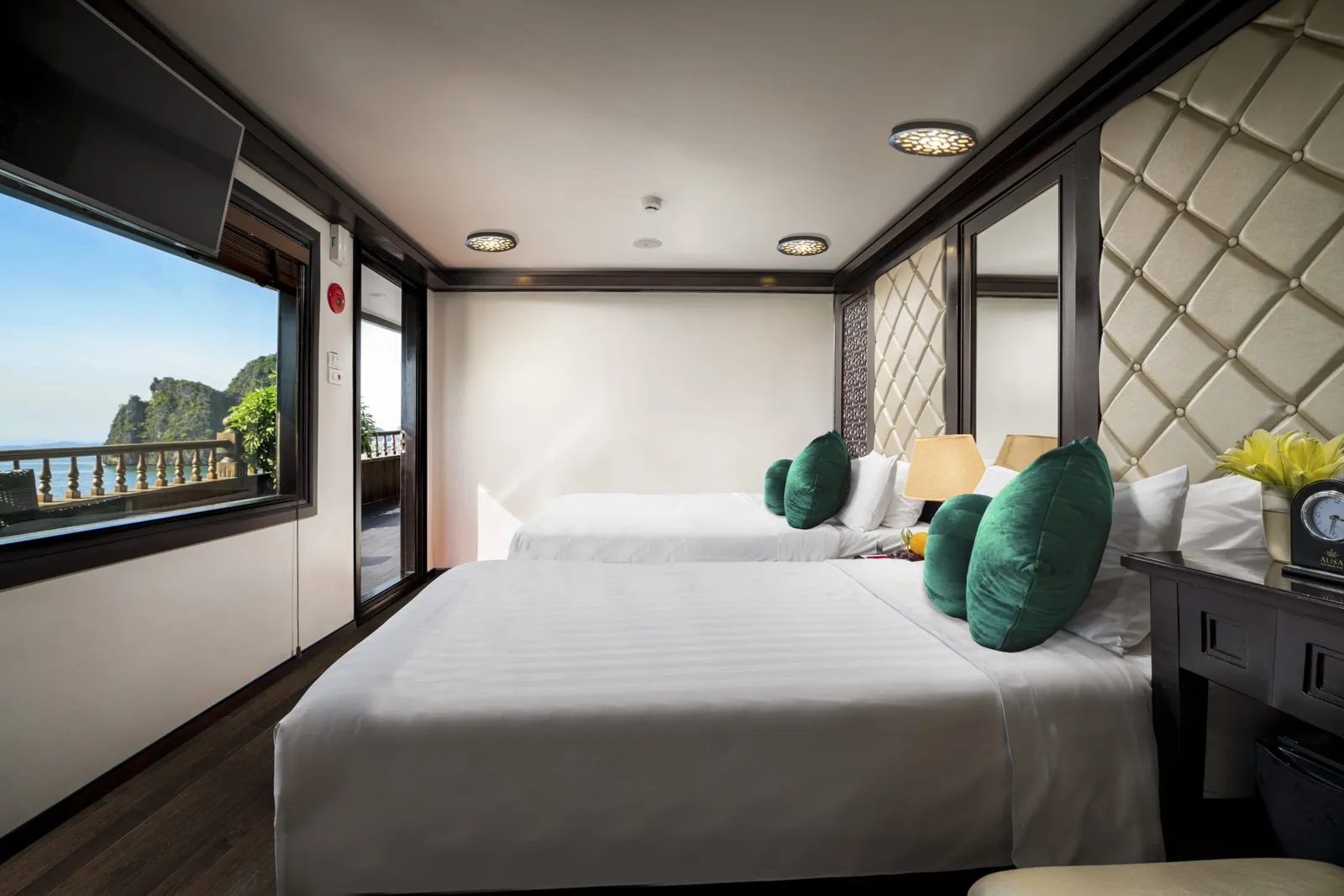 A cruise cabin interior with two beds, green cushions, padded headboard, and windows showing limestone karst formations and water outside.