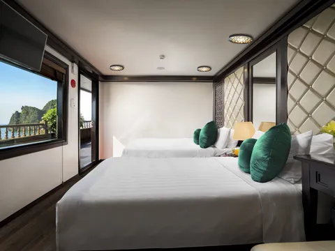 A cruise cabin interior with two beds, green cushions, padded headboard, and windows showing limestone karst formations and water outside.