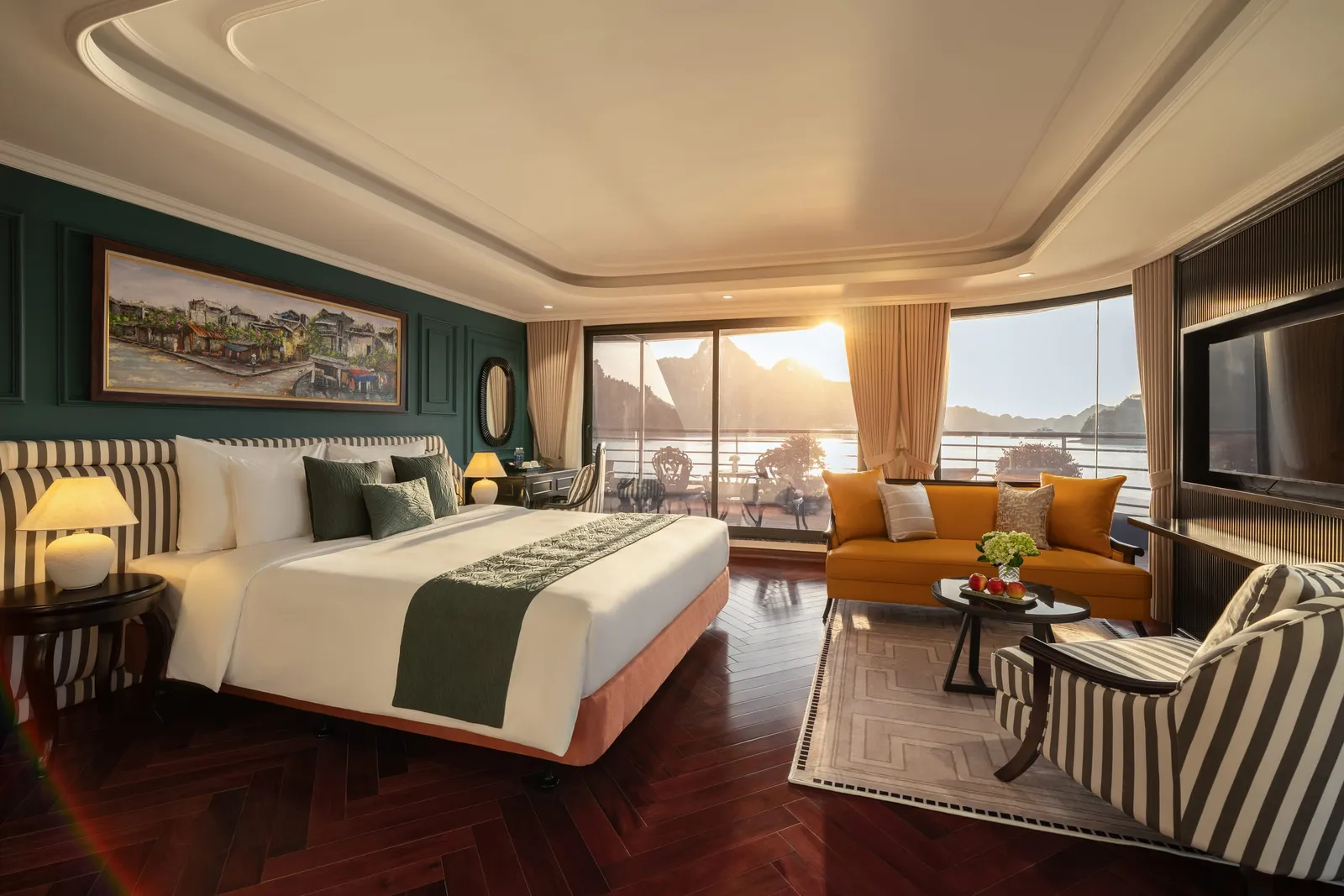 A luxury cabin interior with a bed, orange sofa, and large windows showing karst mountains and water at sunset.