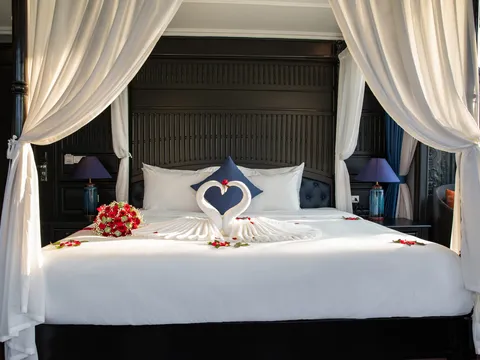 Four-poster bed under white drapes, towels folded into two swans over a heart of red petals, lamps either side