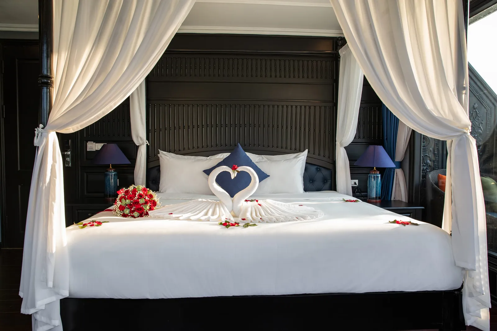 Four-poster bed under white drapes, towels folded into two swans over a heart of red petals, lamps either side