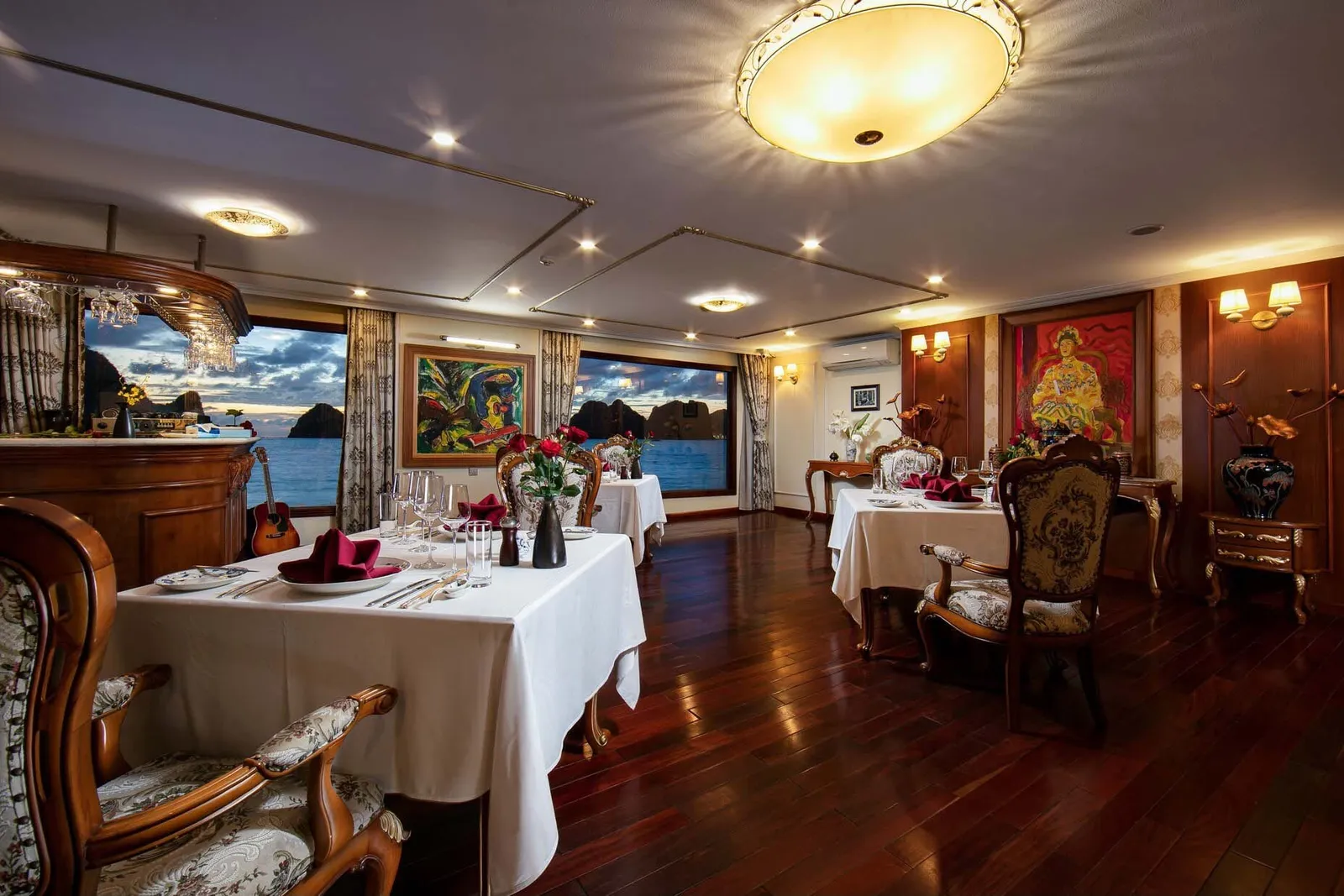 An elegant dining room with white-clothed tables, ornate chairs, decorative artwork, and large windows showing karst limestone islands and water outside.