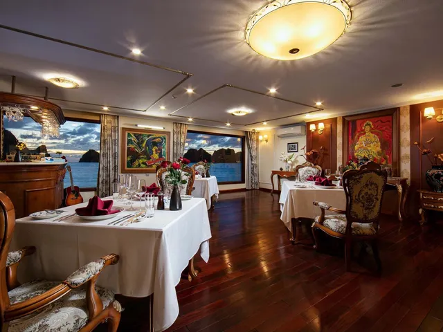 An elegant dining room with white-clothed tables, ornate chairs, decorative artwork, and large windows showing karst limestone islands and water outside.