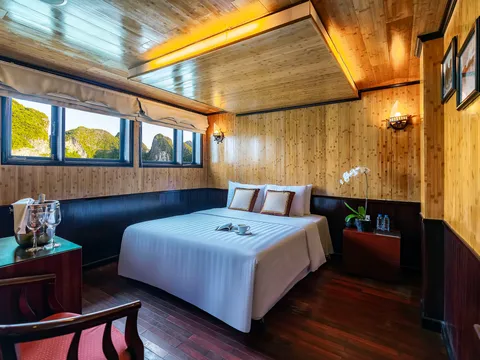 A wood-paneled cabin with a white-draped double bed, wine glasses in an ice bucket, an orchid on a bedside table, and windows showing green limestone karst peaks.