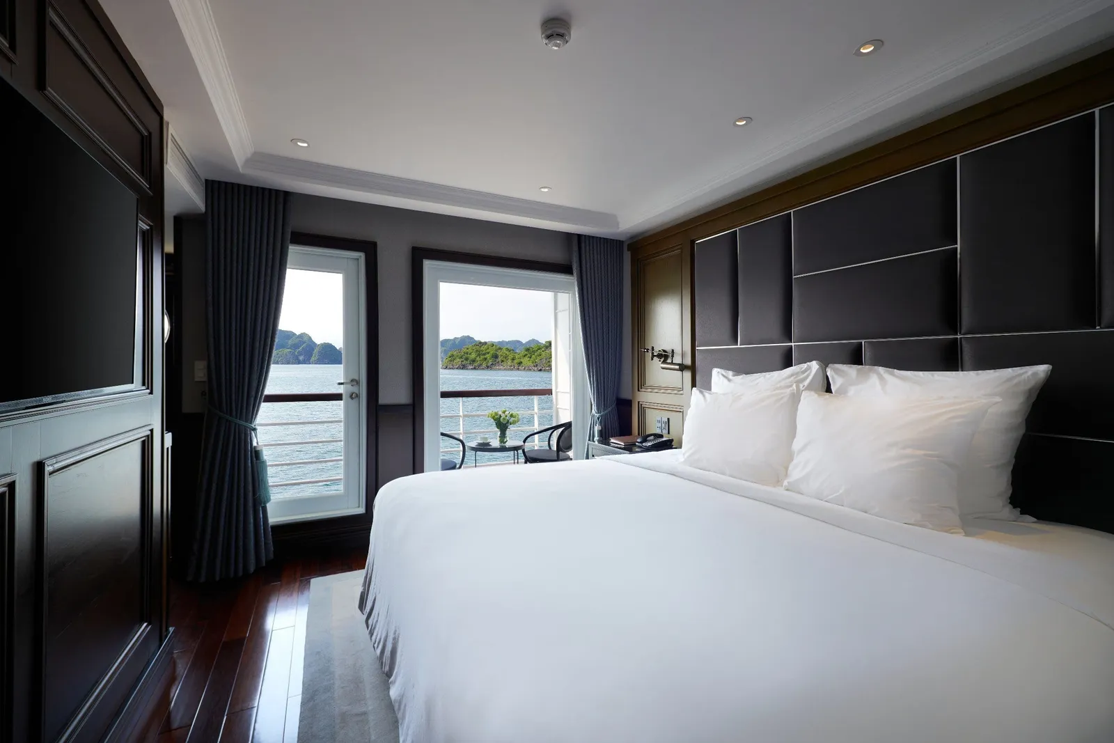 A luxury cabin interior with a white bed, dark wood flooring, and glass doors opening to a balcony with limestone karst islands visible across the water.