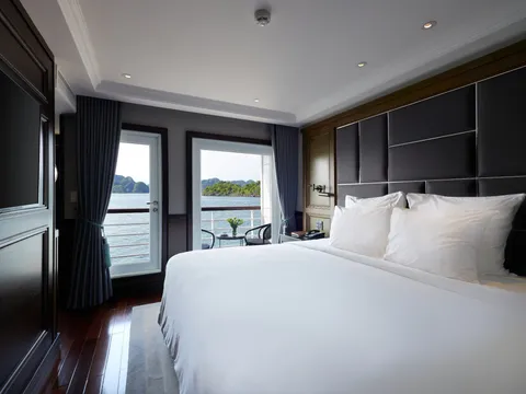 A luxury cabin interior with a white bed, dark wood flooring, and glass doors opening to a balcony with limestone karst islands visible across the water.
