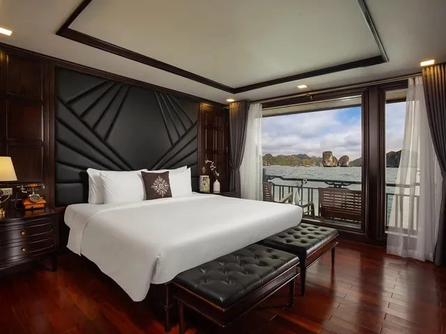 A luxury cabin interior with a white bed, dark wood paneling, and large windows showing limestone karsts and water outside.