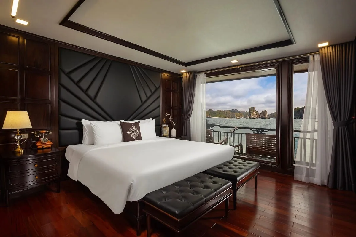 A luxury cabin interior with a white bed, dark wood paneling, and large windows showing limestone karsts and water outside.