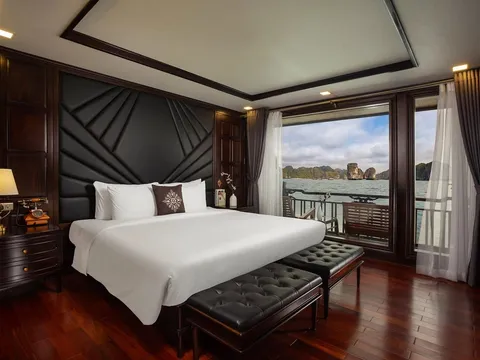 A luxury cabin interior with a white bed, dark wood paneling, and large windows showing limestone karsts and water outside.