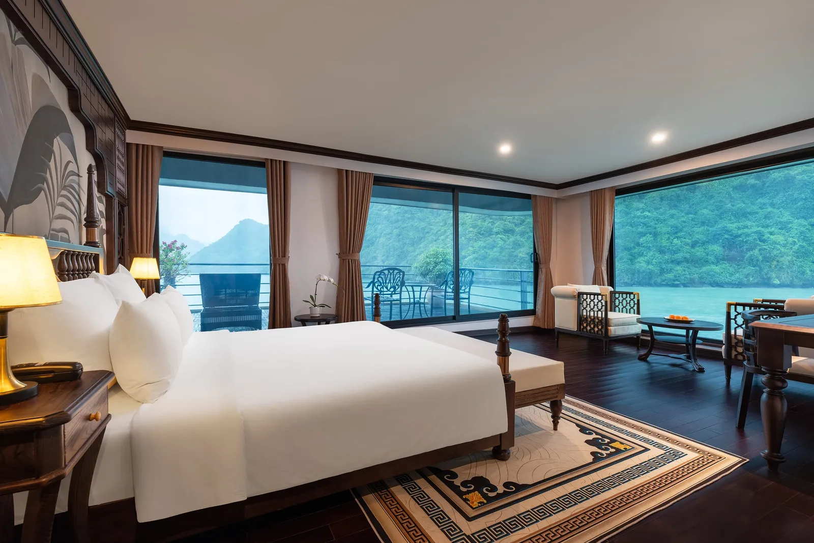 A spacious luxury cabin with a king bed, dark wood furniture, and floor-to-ceiling windows overlooking karst limestone islands on calm green water.