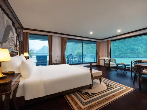 A spacious luxury cabin with a king bed, dark wood furniture, and floor-to-ceiling windows overlooking karst limestone islands on calm green water.