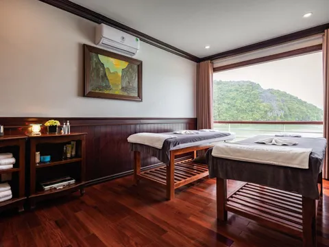 A spa or massage cabin with two treatment tables, dark wood furnishings, and a large window showing a limestone karst island on calm water.