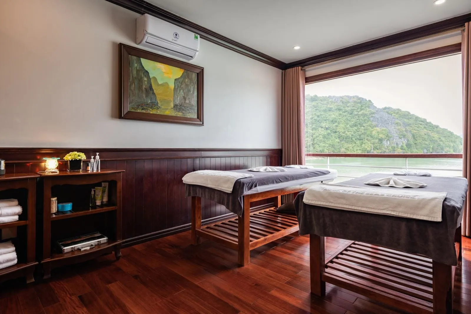 A spa or massage cabin with two treatment tables, dark wood furnishings, and a large window showing a limestone karst island on calm water.