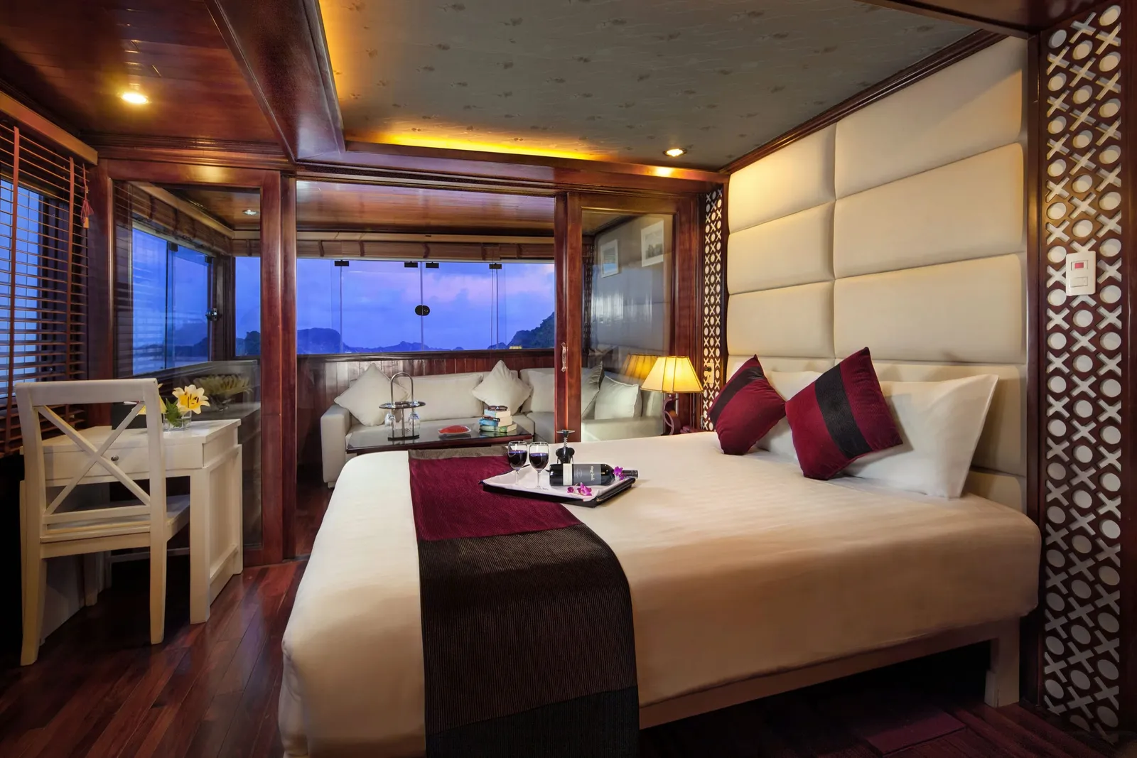 A luxury cabin interior with a white bed, dark wood paneling, and windows showing karst limestone formations at dusk.