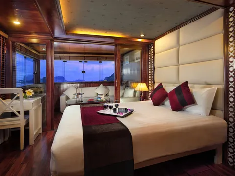 A luxury cabin interior with a white bed, dark wood paneling, and windows showing karst limestone formations at dusk.