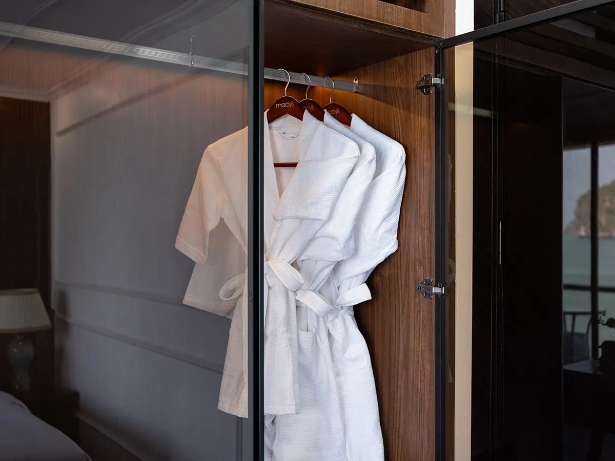Three white waffle bathrobes hang on 'macy's' wooden hangers in an open wardrobe inside a cabin, with a limestone karst island and sea visible through the glass door on the right.
